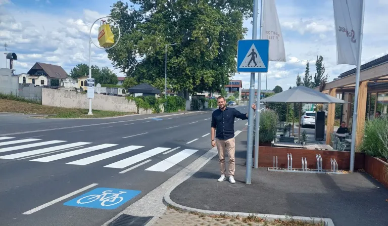 Ein Mann steht neben einem Fußgängerübergangsschild auf einer Straße mit Fahrradweg. Es gibt einen gelben Beutel an einem Pfosten und einen Baum im Hintergrund.