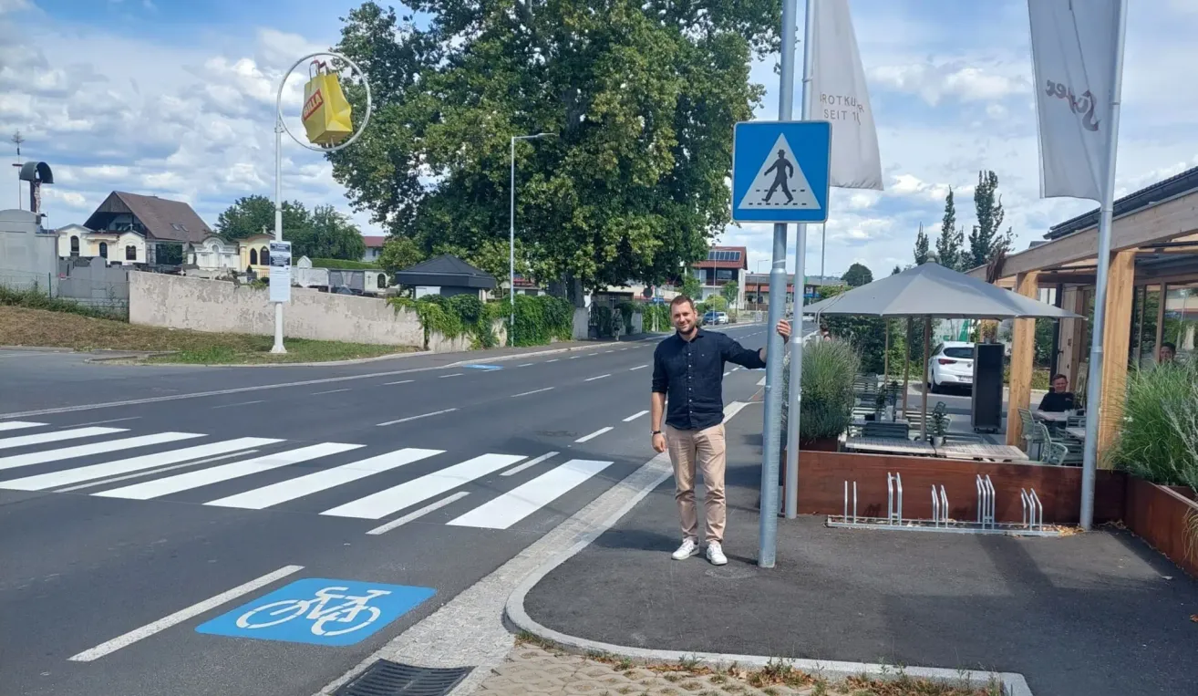 Ein Mann steht neben einem Fußgängerübergangsschild auf einer Straße mit Fahrradweg. Es gibt einen gelben Beutel an einem Pfosten und einen Baum im Hintergrund.