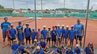 Eine Gruppe von Kindern in blauen Uniformen, einige mit Tennisschlägern, posiert für ein Foto auf einem Tennisplatz mit einem Zaun im Hintergrund.