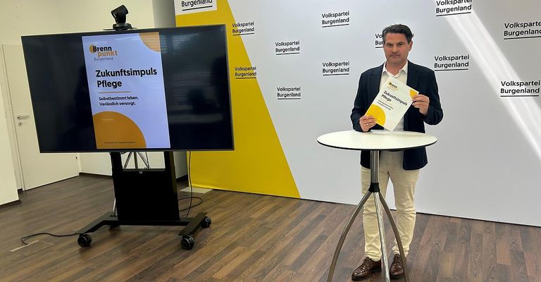 Ein Mann steht vor einem Tisch und hält ein Buch mit dem Titel Zukunftsimpuls Pflege in der Hand, mit einem großen Bildschirm dahinter, der einen ähnlichen Titel anzeigt.