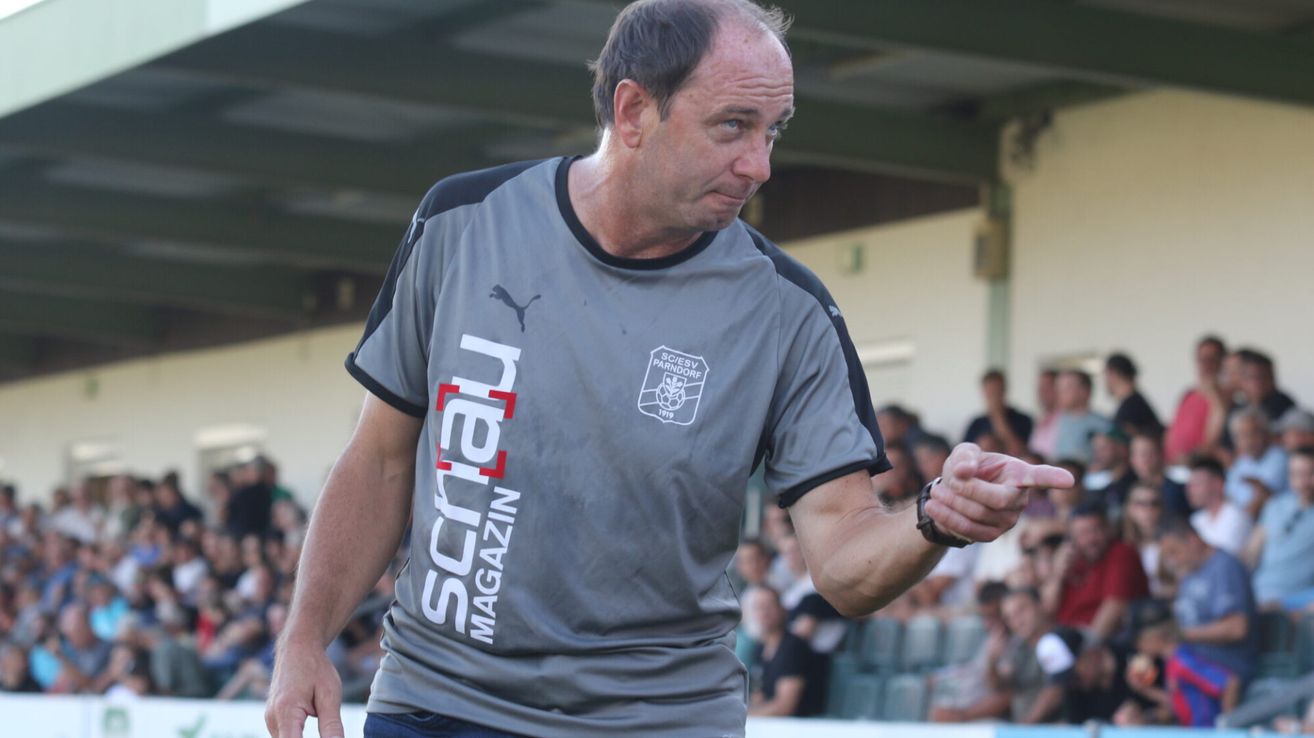 Ein Mann in einem grauen Puma-Shirt mit der Aufschrift 'SCRTAL MAGAZIN' macht eine Geste mit der Hand. Er steht vor einer Menge.