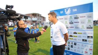 Ein Mann in einem weißen T-Shirt wird von einem Reporter auf einem Fußballfeld interviewt. Hinter ihm ist ein blauer Banner mit Logos und dem Jahr 1946 zu sehen.