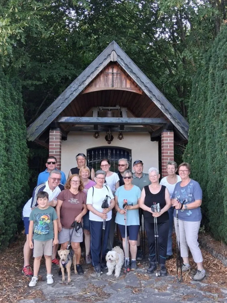 Eine Gruppe von Menschen, einige mit Wanderstöcken, steht vor einer kleinen Holzkapelle mit einem Kreuz auf dem Dach. Sie lächeln und scheinen für ein Foto zu posieren.