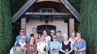 Eine Gruppe von Menschen, einige mit Wanderstöcken, steht vor einer kleinen Holzkapelle mit einem Kreuz auf dem Dach. Sie lächeln und scheinen für ein Foto zu posieren.