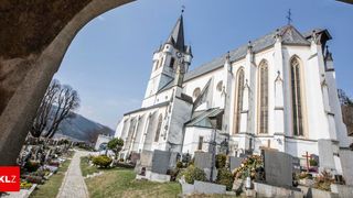 Eine weiße Kirche mit einem Turm und Uhren steht in einem Friedhof mit Grabsteinen und einem Weg unter einem klaren blauen Himmel.