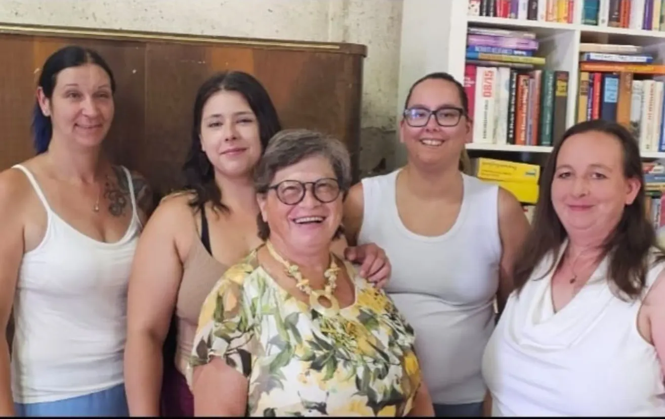 Vier Frauen lächeln und posieren für ein Foto in einem Raum mit einem Bücherregal im Hintergrund.