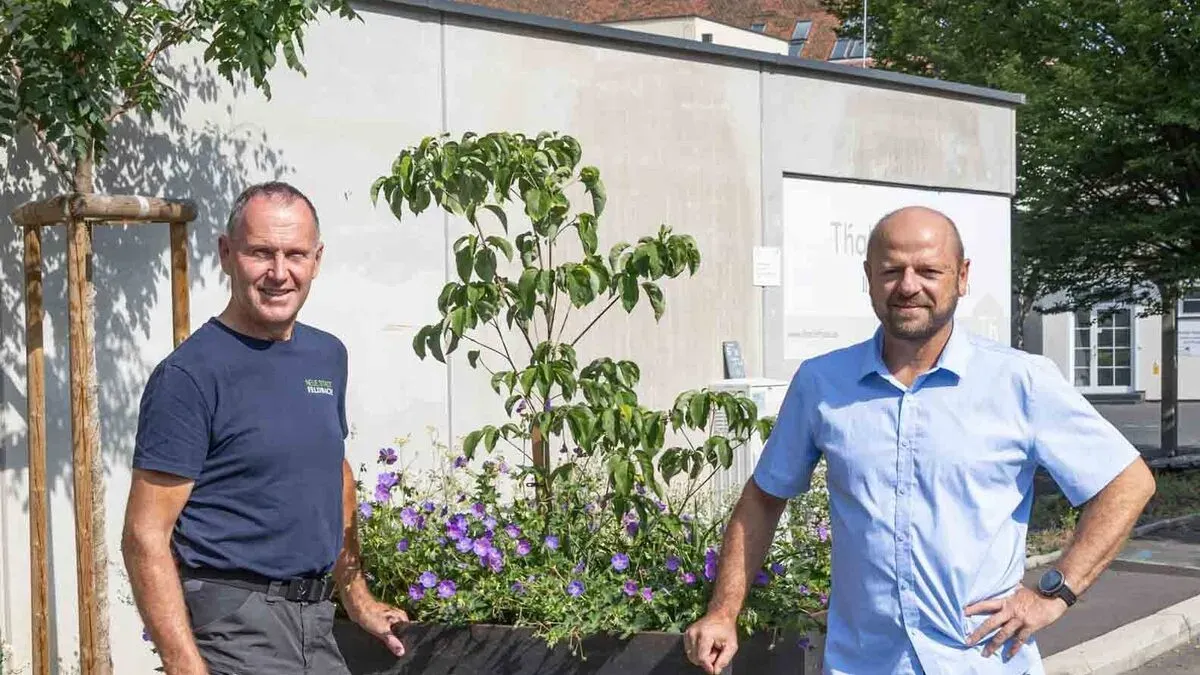 Zwei Männer stehen neben einem Pflanzgefäß mit einem kleinen Baum und lila Blumen und lächeln in die Kamera. Der Mann auf der linken Seite trägt ein blaues Shirt mit weißem Text, während der Mann auf der rechten Seite ein hellblaues Shirt trägt. Hinter ihnen befindet sich eine weiße Wand mit einer weißen Tür und einem Schild, auf dem 'The' steht.