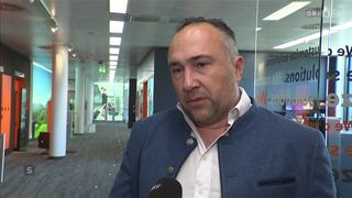 Ein Mann in einem blauen Jackett und weißem Hemd wird in einem Büroflur interviewt. Hinter ihm spiegelt eine Glaswand einen Korridor mit einer Person wider.