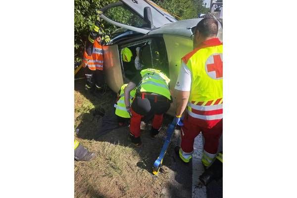 Rettungskräfte in gelben Westen kümmern sich um eine Person in einem beschädigten Fahrzeug. Ein Helfer benutzt ein Werkzeug, um der Person zu helfen.
