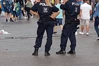 Zwei Polizeibeamte in Uniform stehen inmitten einer belebten Straße, um sie herum gehen Menschen.