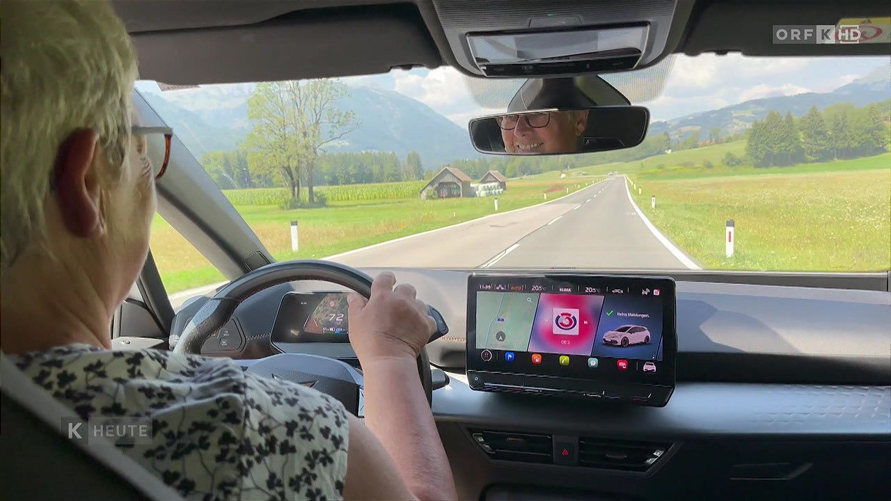 Eine Person, die ein Auto fährt, mit Blick auf die Straße, Berge und ein Haus in der Ferne. Das Armaturenbrett zeigt einen Navigationsbildschirm und ein Tachometer.