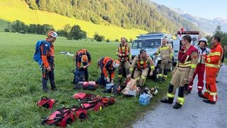Eine Gruppe von Rettungskräften in orange und gelben Uniformen bereitet medizinische Ausrüstung an einem Grasweg vor, mit Bergen im Hintergrund.