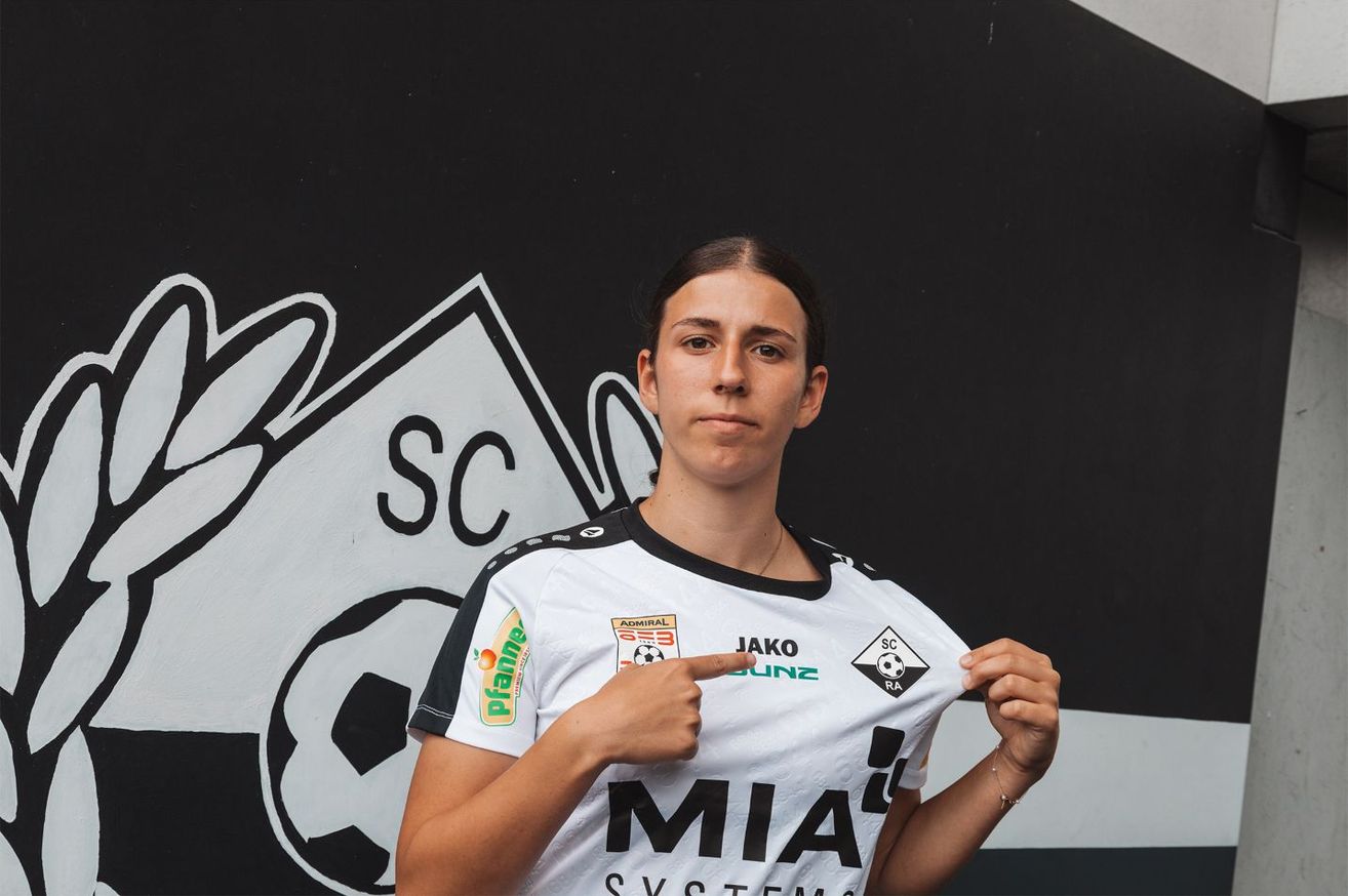 Eine Frau in einem weißen Fußballtrikot mit verschiedenen Logos zeigt auf ihr Shirt, während sie vor einer Graffitiwand steht.
