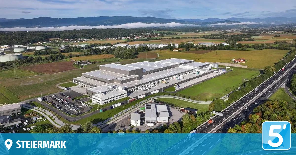 Eine Luftaufnahme eines großen Industriekomplexes mit mehreren Gebäuden, einem Parkplatz und einer Autobahn. Der Komplex ist von grünen Feldern und Bäumen umgeben.