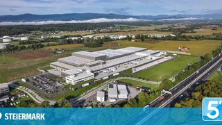 Eine Luftaufnahme eines großen Industriekomplexes mit mehreren Gebäuden, einem Parkplatz und einer Autobahn. Der Komplex ist von grünen Feldern und Bäumen umgeben.