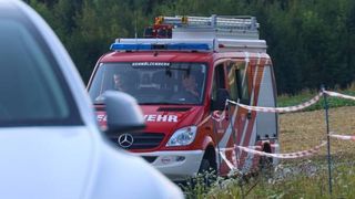 Ein rotes Notfallfahrzeug mit dem Wort 'Feuerwehr' an der Seite ist am Straßenrand geparkt, mit einem weißen Auto davor.