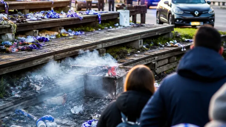 Eine rauchige Szene mit einer Person, die auf einem Grill Essen brät, umgeben von Trümmern und Zuschauern.