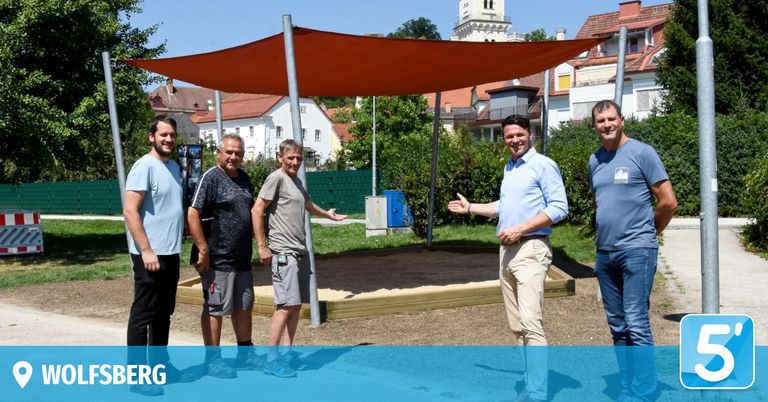 Fünf Männer stehen in einem Park mit einem großen orangefarbenen Baldachin. Sie lächeln und scheinen für ein Foto zu posieren. Hinter ihnen befindet sich ein Sandkasten mit Holzrändern.