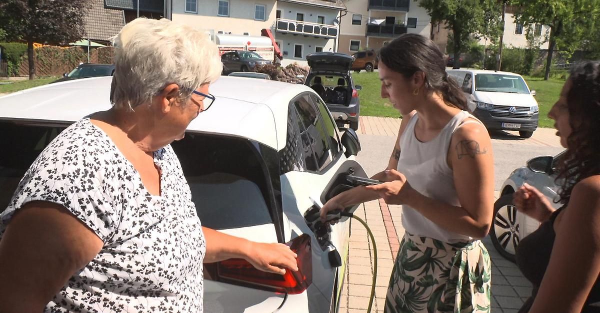 Eine ältere Frau mit Brille hilft einer jüngeren Frau, ein weißes Elektroauto an einer Ladestation am Straßenrand anzuschließen.