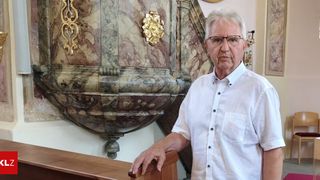 Ein älterer Mann mit Brille und weißem Hemd steht vor einer marmornen Kanzel mit goldenen Verzierungen.
