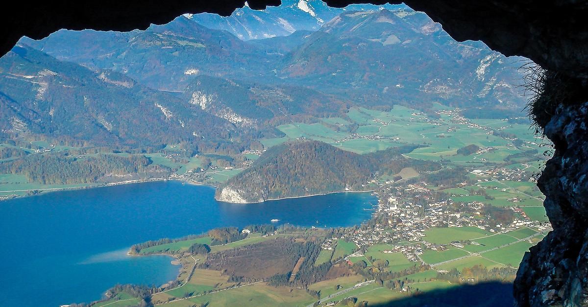 Luftaufnahme eines ruhigen Sees, umgeben von üppigen grünen Feldern und dichten Wäldern, mit einem kleinen Dorf am Seeufer und majestätischen Bergen im Hintergrund.
