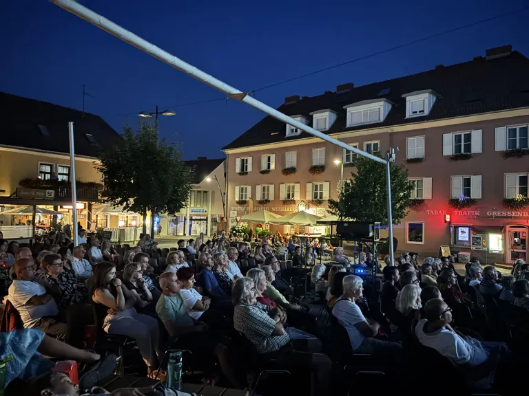 Eine Menschenmenge sitzt in Stühlen unter einem Zelt in einer Freiluftumgebung bei Nacht. Das Publikum sieht einer Bühne mit einem Lautsprecher und einem Bildschirm zu. Im Hintergrund befinden sich Gebäude mit Fenstern und Straßenlaternen.