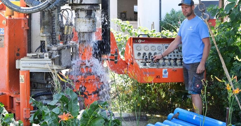 Ein Mann bedient eine große orange Bohrmaschine in einem Garten und spritzt Wasser, während sie bohrt.
