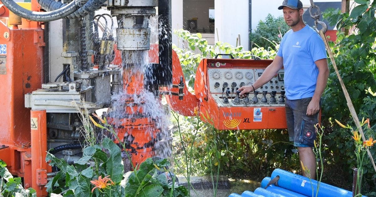 Ein Mann bedient eine große orange Bohrmaschine in einem Garten und spritzt Wasser, während sie bohrt.