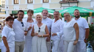 Eine Gruppe von Menschen in weißen Kleidern steht draußen, lächelt und hält Weingläser in der Hand. Sie scheinen bei einer sozialen Veranstaltung zu sein.