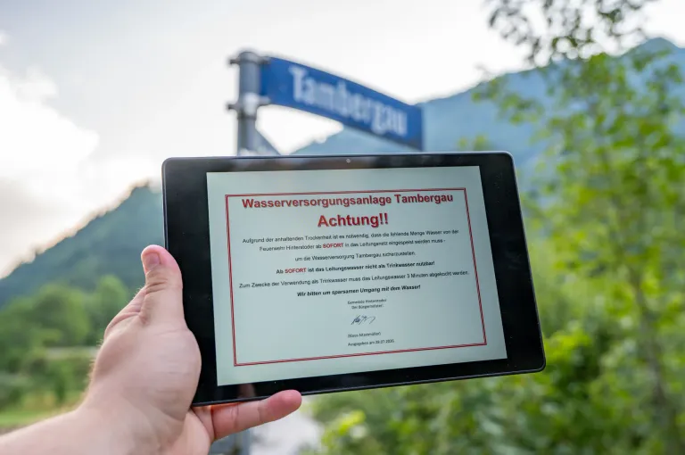 Eine Hand hält ein Tablet, das einen Warnhinweis in deutscher Sprache über Wasserversorgungsprobleme in Tambergau anzeigt. Der Hinweis rät, Leitungswasser drei Minuten lang abzukochen, bevor es getrunken wird.