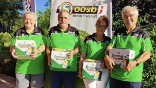 Vier Personen in grünen Polo-Shirts stehen zusammen, jeder hält eine Kiste Freistadter Bier. Der Mann in der Mitte hat einen Schnurrbart und trägt eine Uhr. Hinter ihnen ist ein Banner mit den Worten 'Bezirk Freistadt'.