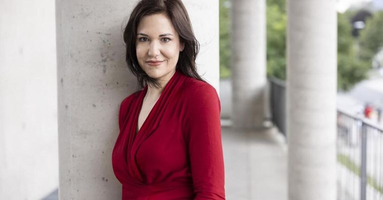 Eine Frau mit dunklem Haar, die ein rotes Kleid trägt, steht lächelnd vor einer Betonwand mit Säulen.