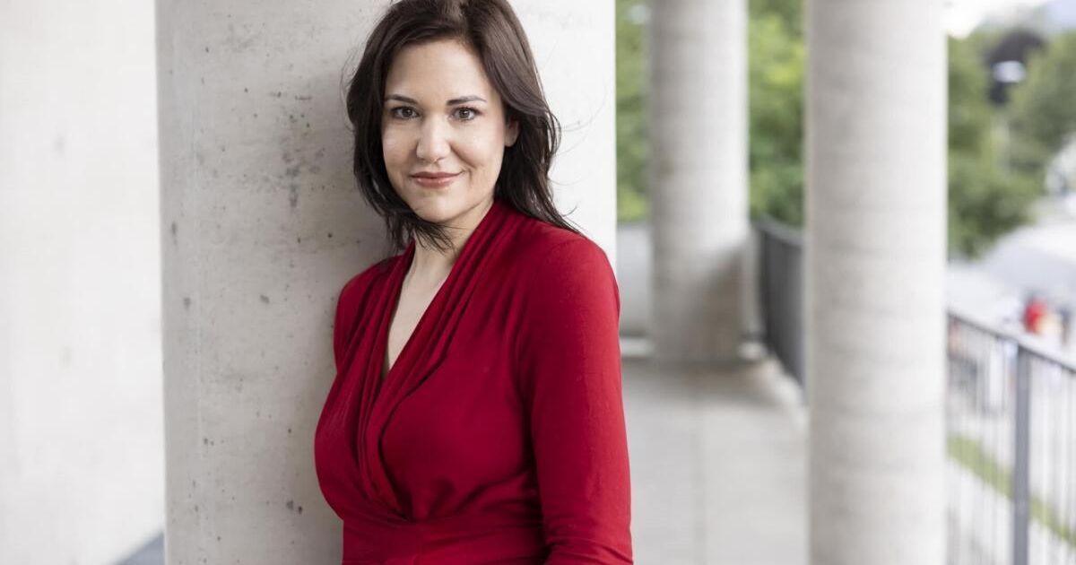 Eine Frau mit dunklem Haar, die ein rotes Kleid trägt, steht lächelnd vor einer Betonwand mit Säulen.