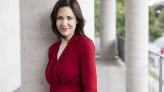 Eine Frau mit dunklem Haar, die ein rotes Kleid trägt, steht lächelnd vor einer Betonwand mit Säulen.