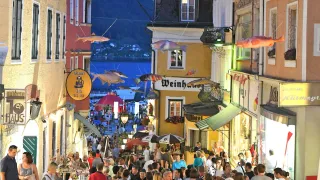 Eine überfüllte Straße in einer europäischen Stadt bei Nacht, mit großen Fischdekorationen darüber und Geschäften auf beiden Seiten. Menschen gehen vorbei, einige tragen Taschen. In der Ferne sind Berge und ein Gewässer zu sehen.