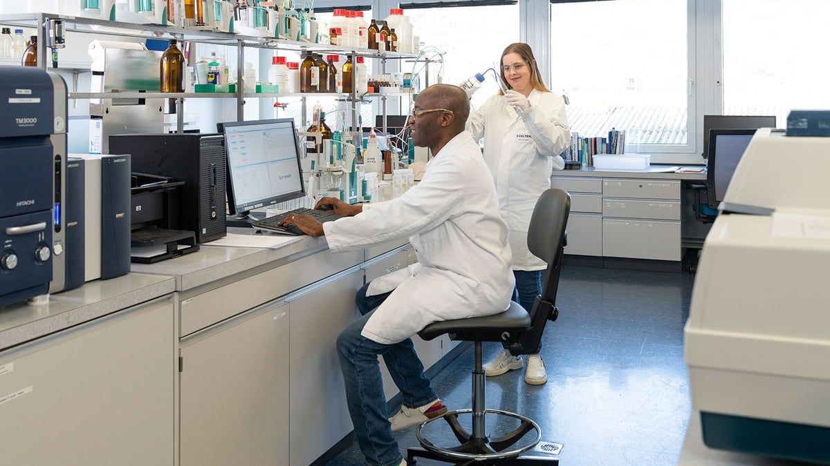 Zwei Wissenschaftler in weißen Laborkittel arbeiten in einem Labor. Einer tippt auf einem Computer, während der andere eine Pipette hält. Hinter ihnen befinden sich Regale mit Flaschen und Geräten.