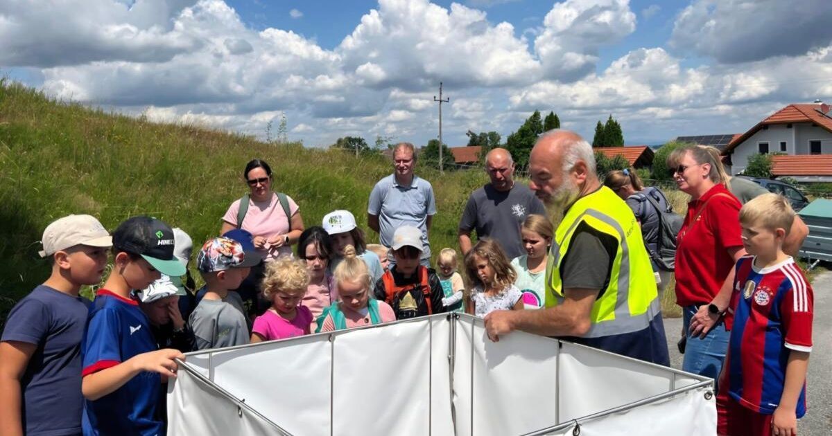 Eine Gruppe von Kindern und Erwachsenen versammelt sich um ein weißes Zelt auf einer Wiese, wobei ein Mann in einer Warnweste ihnen etwas erklärt.