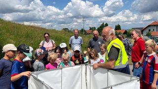 Eine Gruppe von Kindern und Erwachsenen versammelt sich um ein weißes Zelt auf einer Wiese, wobei ein Mann in einer Warnweste ihnen etwas erklärt.