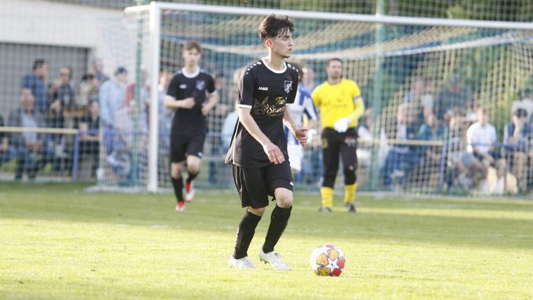 Ein Fußballspieler in einem schwarzen Trikot mit 'JAKO' und 'The Savage' darauf läuft auf einen Fußball auf einem Rasenfeld zu.
