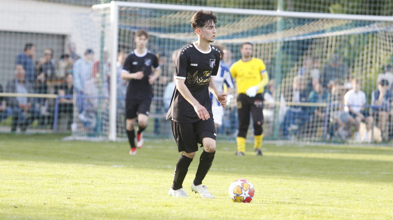 Ein Fußballspieler in einem schwarzen Trikot mit 'JAKO' und 'The Savage' darauf läuft auf einen Fußball auf einem Rasenfeld zu.