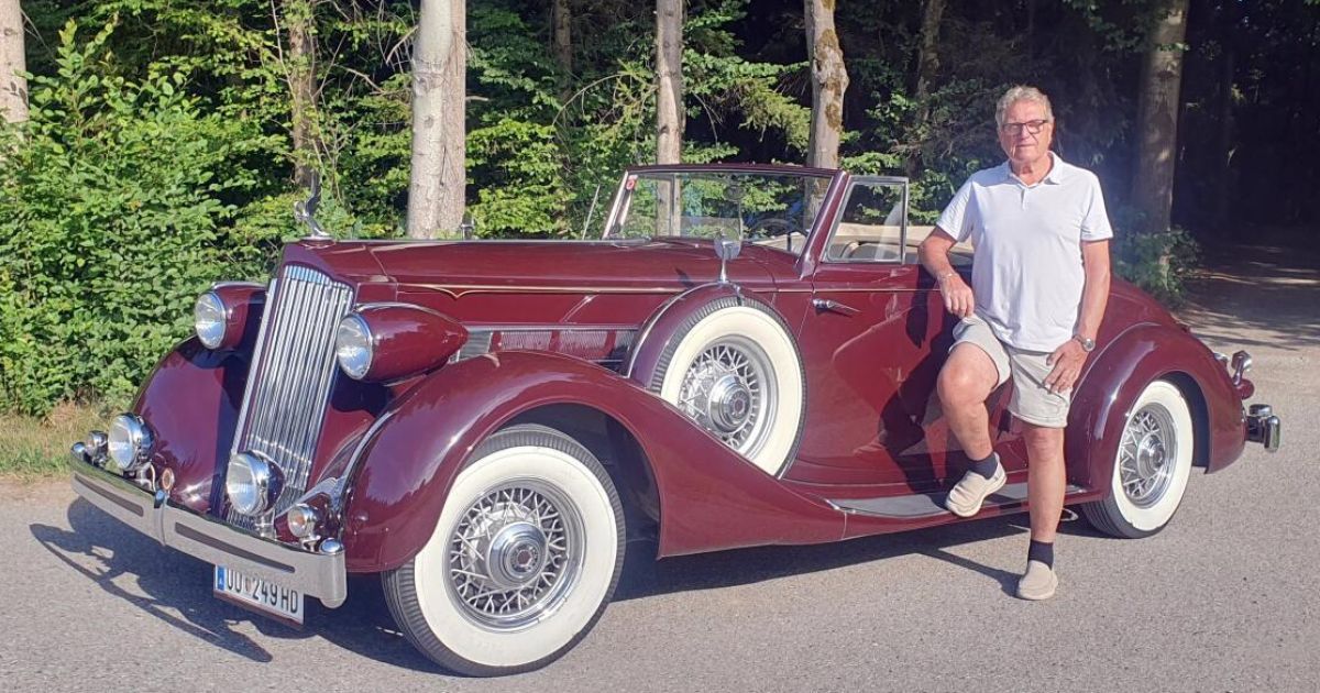 Ein älterer Mann steht neben einem burgunderroten Oldtimer mit weißen Felgen und einem Nummernschild, das 249 HD liest. Er trägt ein weißes Poloshirt, Shorts und weiße Socken. Das Auto ist am Straßenrand geparkt, mit Bäumen im Hintergrund.