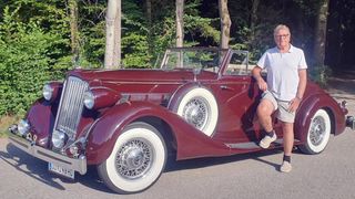 Ein älterer Mann steht neben einem burgunderroten Oldtimer mit weißen Felgen und einem Nummernschild, das 249 HD liest. Er trägt ein weißes Poloshirt, Shorts und weiße Socken. Das Auto ist am Straßenrand geparkt, mit Bäumen im Hintergrund.