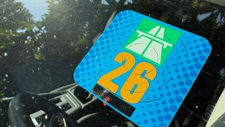 Ein Aufkleber mit der Nummer 26 und einem grünen Logo befindet sich auf der Windschutzscheibe eines Autos. Der Aufkleber hat einen blauen Hintergrund mit blauen Punkten.