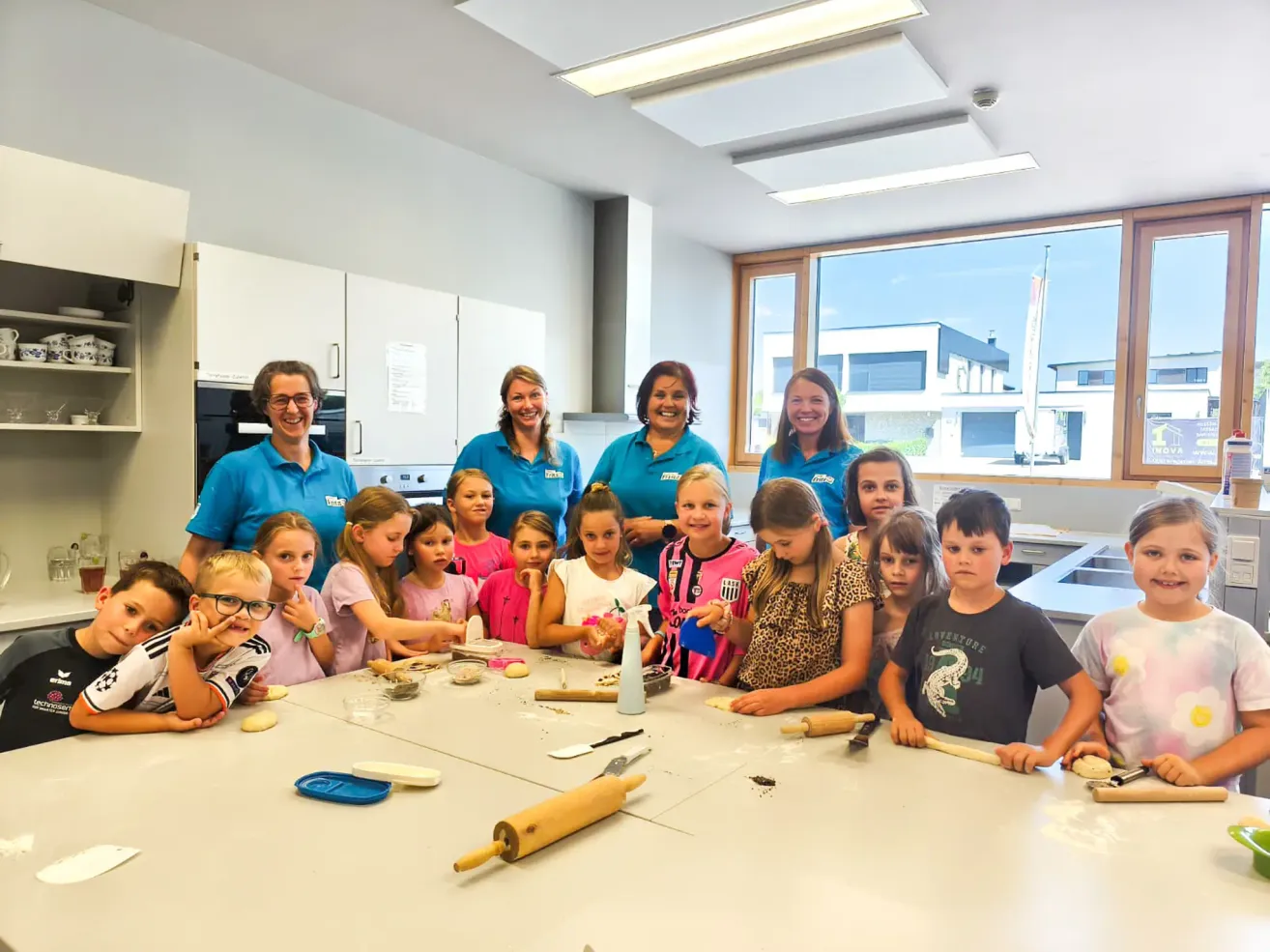 Eine Gruppe von Kindern und Erwachsenen in einer Küche, lächelnd und posierend für ein Foto, während sie Teig herstellen. Die Kinder sitzen um einen Tisch mit Nudelholz und Schüsseln mit Teig. Die Erwachsenen stehen hinter ihnen, lächelnd und beobachtend.
