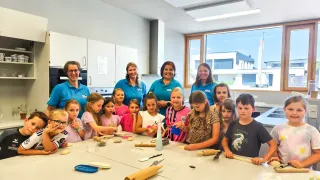 Eine Gruppe von Kindern und Erwachsenen in einer Küche, lächelnd und posierend für ein Foto, während sie Teig herstellen. Die Kinder sitzen um einen Tisch mit Nudelholz und Schüsseln mit Teig. Die Erwachsenen stehen hinter ihnen, lächelnd und beobachtend.