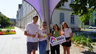 Vier Personen stehen unter einem violetten Baldachin und halten Eis am Stiel. Sie tragen Sonnenbrillen und lächeln. Eine Frau hält ein Schild mit der Aufschrift 'Weiz Deine junge Stadt'. Hinter ihnen steht ein Gebäude mit vielen Fenstern.