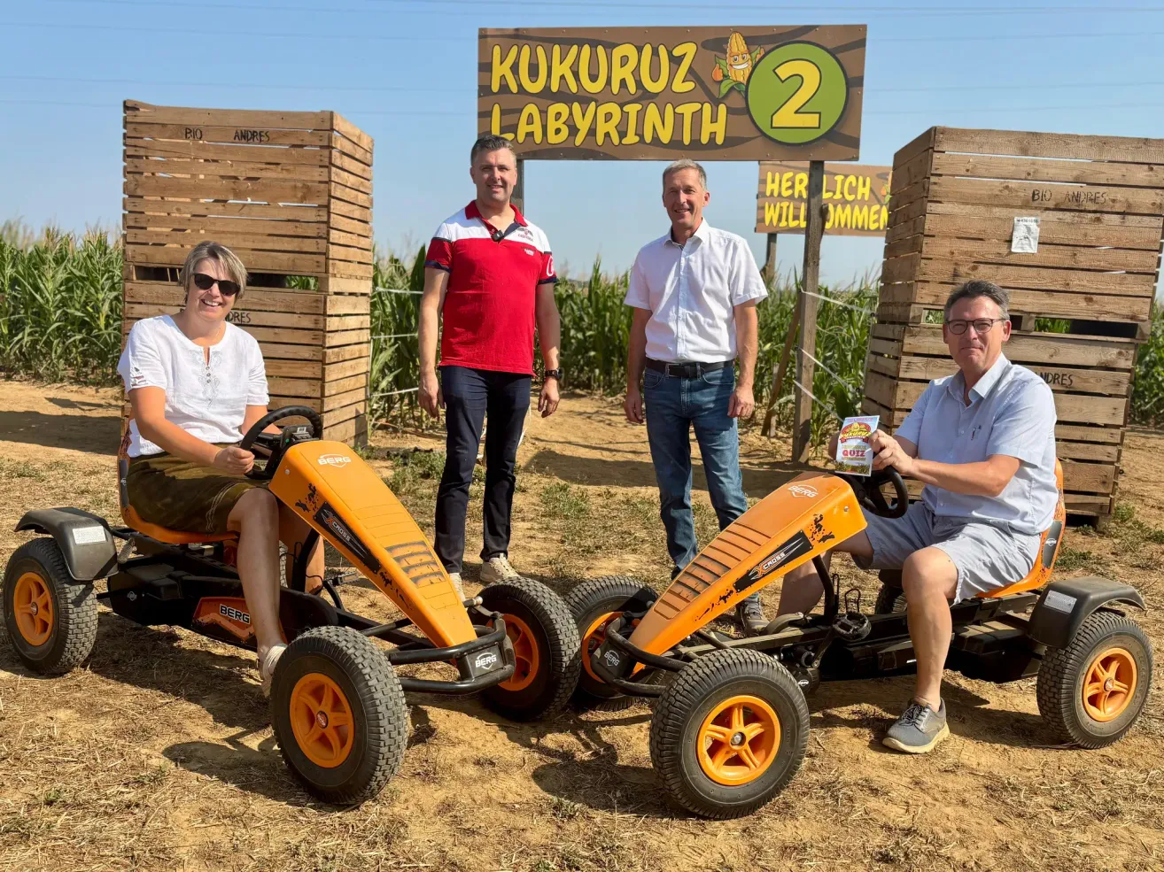 Vier Erwachsene posieren vor einem Holzschild, das 'KUKURUZ LABYRINTH 2' besagt. Zwei sitzen in orangefarbenen Go-Karts, während die anderen hinter ihnen stehen. Die Umgebung ist ein Maisfeld.