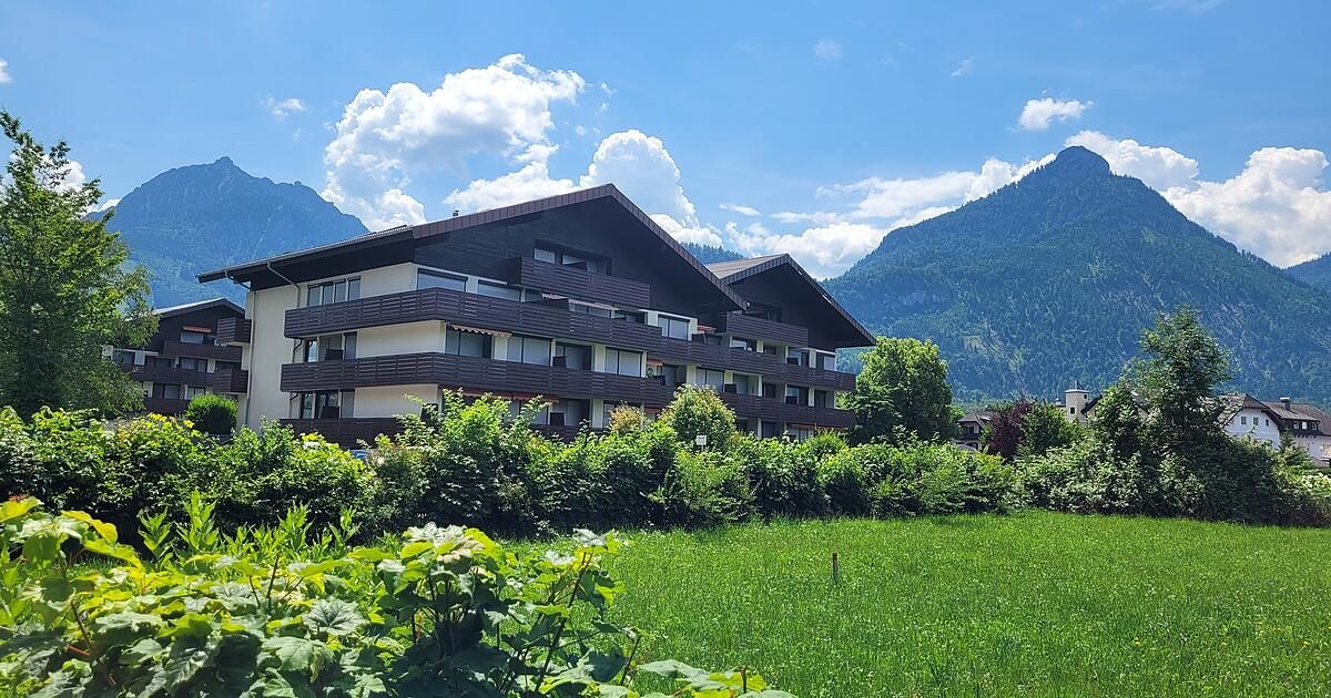 Ein mehrstöckiges Gebäude mit Balkonen und einem grünen Feld davor, vor dem Hintergrund von Bergen und einem blauen Himmel mit Wolken.