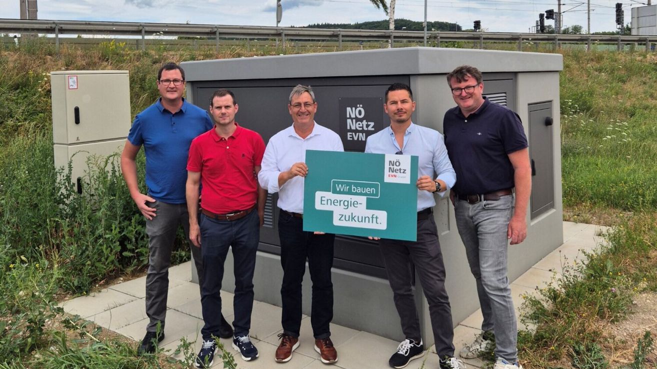 Fünf Männer stehen vor einer grauen Struktur mit einem Schild, auf dem 'NO Netz EVN' steht. Der Mann in der Mitte hält ein grünes Schild mit der Aufschrift 'Wir bauen Energie-zukunft'. Sie alle lächeln und scheinen für ein Foto zu posieren.