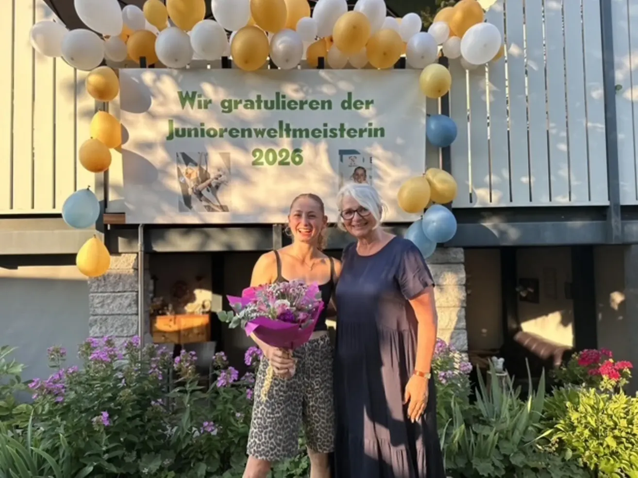Zwei Frauen stehen vor einem Banner mit Luftballons und Blumen, eine hält einen Blumenstrauß und lächelt, die andere trägt eine Brille und lächelt.